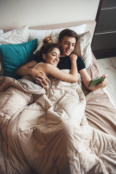 Φωτογραφία του Καυκάσιου ζευγαριού που κείτεται στο κρεβάτι το πρωί και κοιτάζει κάτι στο κινητό ενώ αγκαλιάζει και χαμογελάει. - Φωτογραφία, εικόνα