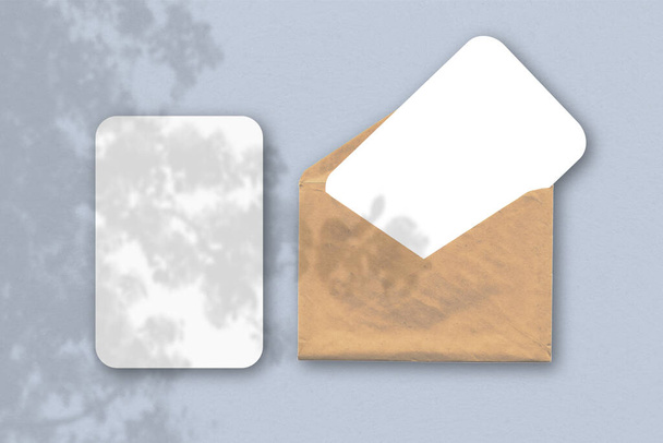 Um envelope com duas folhas de papel branco texturizado sobre um fundo de mesa azul. Mockup com uma sobreposição de sombras de fábrica. A luz natural lança sombras de uma fábrica tropical. Orientação horizontal
 - Foto, Imagem