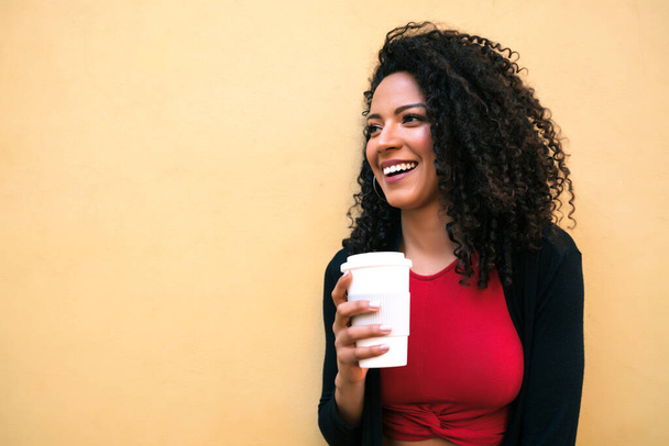 Portrait of young afro american woman looking confident and posing while holding a cup of coffee against yellow background. - Photo, Image