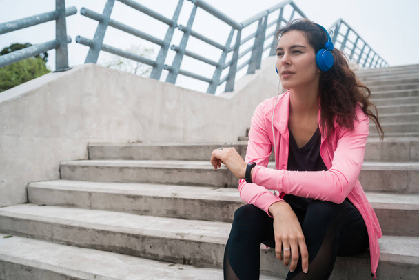 Portrait of an athletic woman listening to music on a break from training while sitting on stairs. Sport and health lifestyle concept. - Фото, зображення