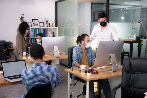 Азиатские офисные работники в масках для лица работают в новом нормальном офисе и делают социальное дистанцирование во время пандемии вируса короны ковид-19 - Фото, изображение
