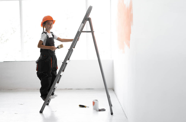 ビルダーの服を着た女の子と頭にオレンジのハードハット。階段を登れ。手にブラシを持っている。ホームリニューアルコンセプト。テキストの場所. - 写真・画像