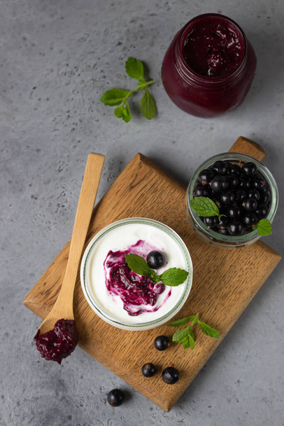 Jar of delicious homemade black currant curd, custard or jam with natural yogurt and fresh berries on grey concrete background. Healthy breakfast. - Fotó, kép