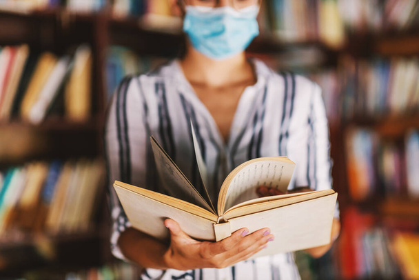 Молода дівчина коледжу з маскою обличчя стоїть в бібліотеці і тримає книгу під час пандемії корони
. - Фото, зображення