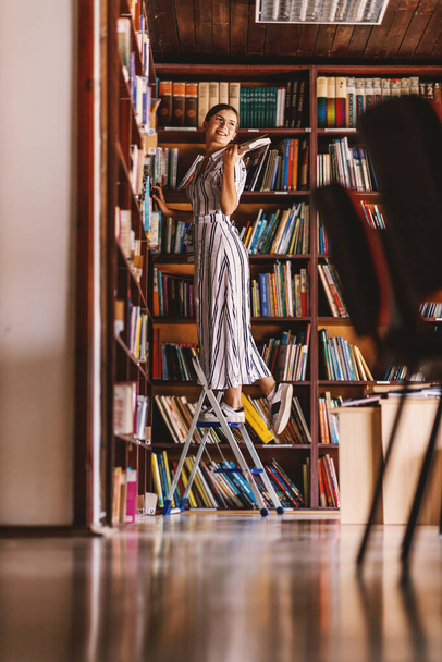 Genç ve çekici kütüphanecinin merdivende durup elinde kitap tutması.. - Fotoğraf, Görsel