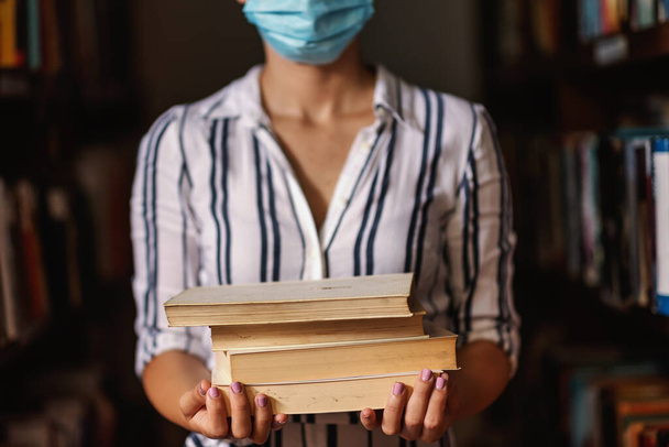 Kütüphanede elinde kitaplarla duran maskeli bir kız. Salgın konsepti sırasında çalışmak. - Fotoğraf, Görsel
