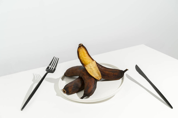 Waste food concept. Stale ugly bananas in a white plate and a black knife and fork on a white background. - Photo, Image