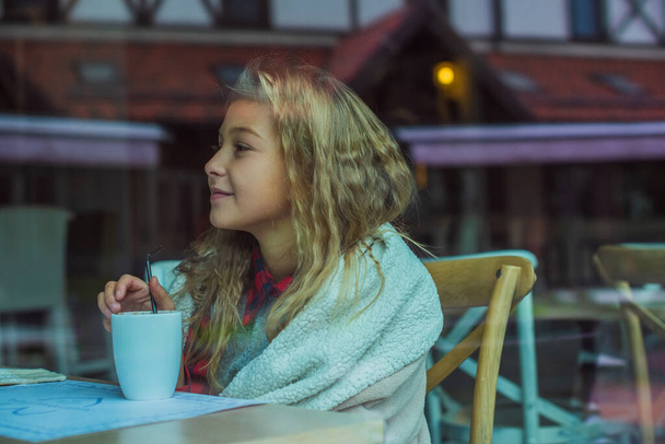 Bonne petite fille au café.L'enfant s'assoit à une table dans un café et est heureux.Une enfance heureuse, concept de confort et d'enfants, saison froide, vacances - Photo, image