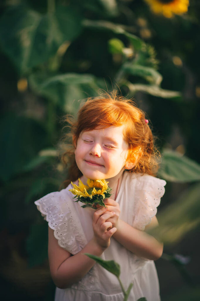 Παιδί σε ένα χωράφι με ηλιοτρόπια. Η κοκκινομάλλα έκλεισε τα μάτια της και έκανε μια ευχή με κλειστά μάτια. Πορτραίτο κλεισίματος - Φωτογραφία, εικόνα