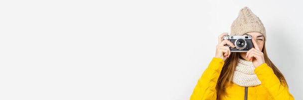 Jonge vrouw in een geel donsjas en hoed houdt een camera in haar handen en maakt een foto op een lichte achtergrond. Concept van schoonheid en mode, winter, herfst, technologie. Banner. - Foto, afbeelding