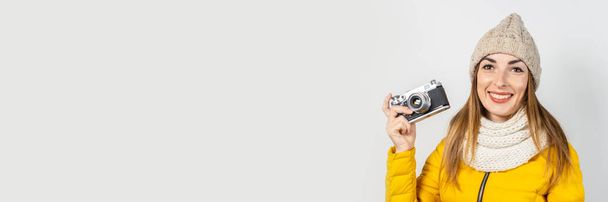 Young woman with smile in a yellow down jacket and hat holds a camera in her hands on a light background. Concept of beauty and fashion, winter, autumn, technology. Banner. - Foto, Imagem