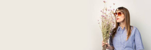 Девушка в солнечных очках держит букет полевых цветов на светлом фоне. Баннер. лето, отпуск, путешествия, косметология. - Фото, изображение