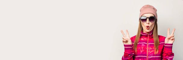 Young woman in glasses, hat and pink sports jacket with a surprised face, makes a cool gesture, in front of white background with copy space. Concept modern trendy style. Face expression. Banner. - Photo, Image