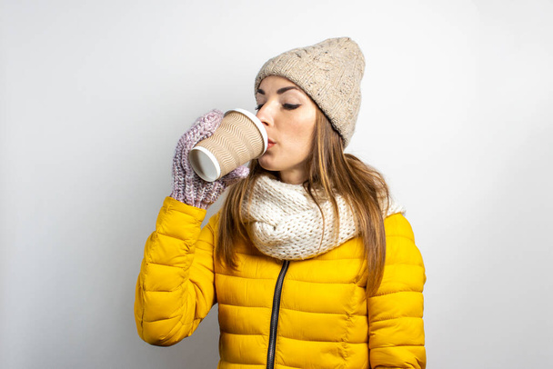 Young woman in a yellow jacket and hat holds a glass of coffee or tea on a light background. Emotion laughter, surprise, kiss. Concept winter, autumn, coffee house. - 写真・画像