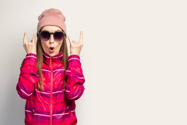 Mujer joven con sombrero y gafas y una chaqueta deportiva rosa hace un gesto de cabra rock and roll, vamos a coger esta fiesta en un fondo blanco con el espacio vacío. Concepto. Expresión facial. Banner. - Foto, Imagen