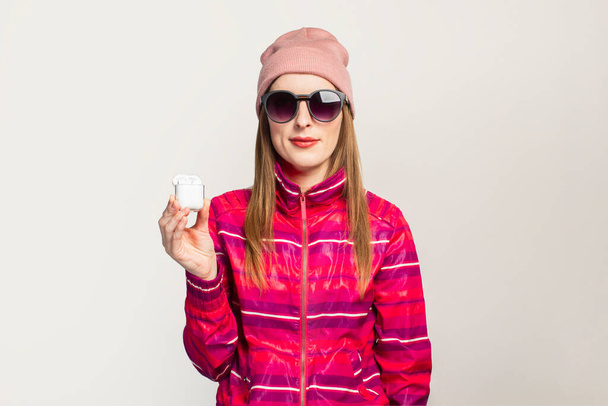 Vriendelijke Jonge vrouw in bril, hoed en roze sportjasje met een smiley gezicht houdt draadloze hoofdtelefoon op een witte achtergrond. Concept moderne stijl, coole muziek. Gezichtsuitdrukking. Banner. - Foto, afbeelding