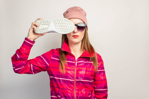 Junge Frau mit Brille, Hut und pinkfarbener Sportjacke mit ernstem Gesicht verdeckt ihr Gesicht mit einem Turnschuh auf weißem Hintergrund mit Kopierraum. Konzept modernen trendigen Stil. Gesichtsausdruck. Banner. - Foto, Bild