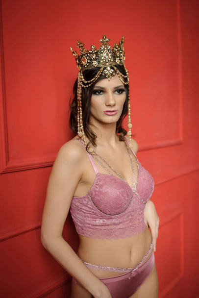 close up photo of a sexy fashion woman in pink underwear with crown on her head on a red background in a photo studio - Photo, Image