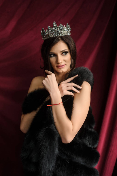 close up photo of a sexy fashion woman in black fur coat with a crown on her head on a red curtain background in a photo studio - Photo, Image