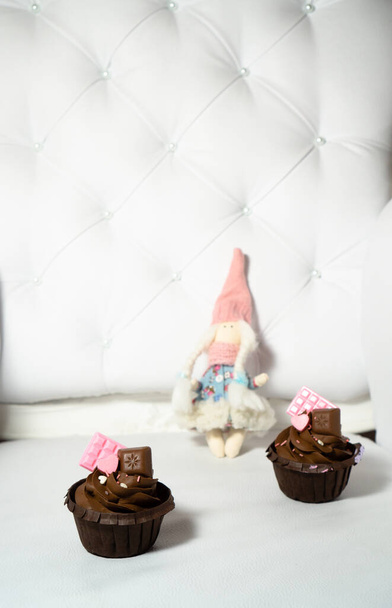 Brązowe babeczki ozdobione są różową czekoladą na lekkim, zabytkowym skórzanym fotelu i lalką w zimowym stroju w tle. - Zdjęcie, obraz