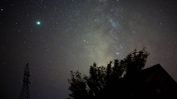 Cielo nocturno estrellado y la Vía Láctea sobre el fondo de un árbol y el techo de una casa por la noche. - Foto, imagen