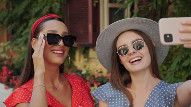 Deux meilleurs amis dans des lunettes de soleil et un chapeau s'amusent, rient, prennent des selfies sur le fond d'une belle maison avec des fleurs dans la ville. - Séquence, vidéo