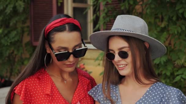Two best cute friends in a sunglasses and hat have fun, laugh, take selfies and look to the pictures on the background of a beautiful house with flowers in the city. - Footage, Video