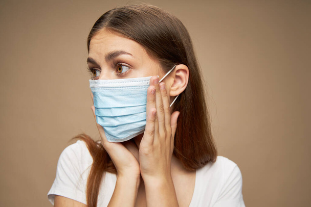 Una mujer con una máscara médica mira a un lado y se toca la cara con las manos sobre un fondo beige  - Foto, imagen
