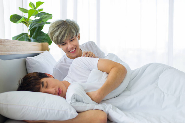 Ευτυχισμένοι Ασιάτες ομοφυλόφιλοι ή γκέι ζευγάρια κείτονται στο λευκό κρεβάτι ενώ ο άλλος προκαλεί τους ώμους του. Έννοια της υπερηφάνειας ΛΟΑΤΚΙ. - Φωτογραφία, εικόνα