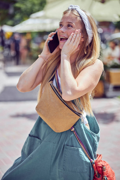 Vidám gyönyörű lány szeplős beszél az okostelefonon, miközben tartja a kezét az arcon oldalnézetet lövés a szabadban   - Fotó, kép