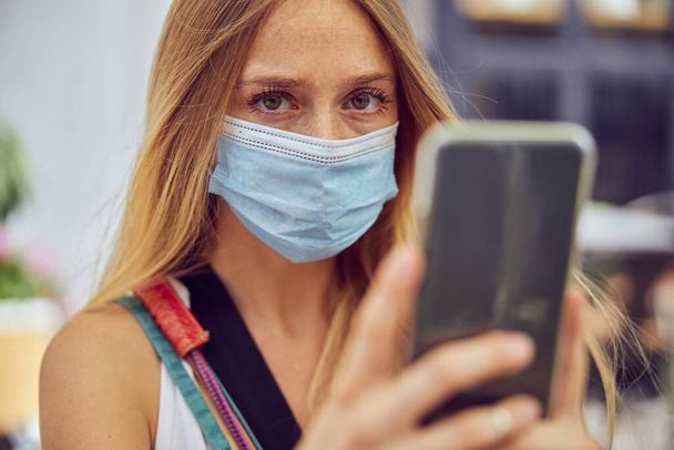 Close up shot of a young girl with freckles wearing a face mask is holding a smartphone while looking at the camera - Photo, Image