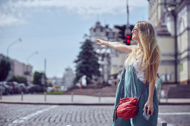   Menina alegre com belos cabelos longos em óculos de sol com smartphone está parando um táxi e mantendo o sorriso perto de passarela uma vista lateral
 - Foto, Imagem