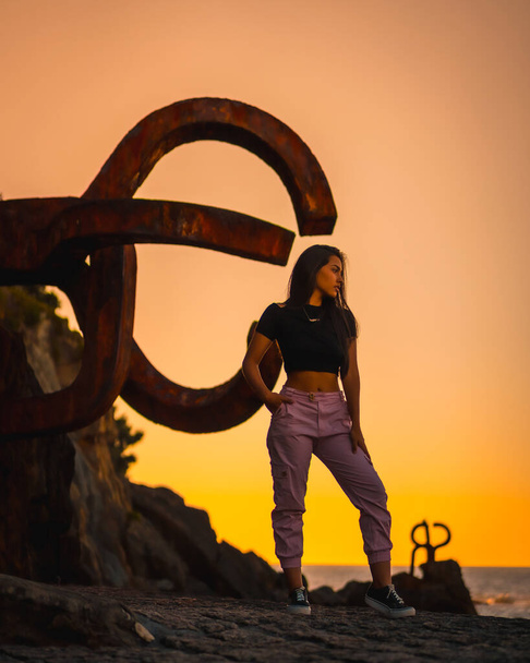 A young pretty brunette Latina with long straight hair in a short black T-shirt and pink pants. Orange sunset next to a famous sculpture of San Sebatian called Peine del Viento, Spain - Foto, Imagem