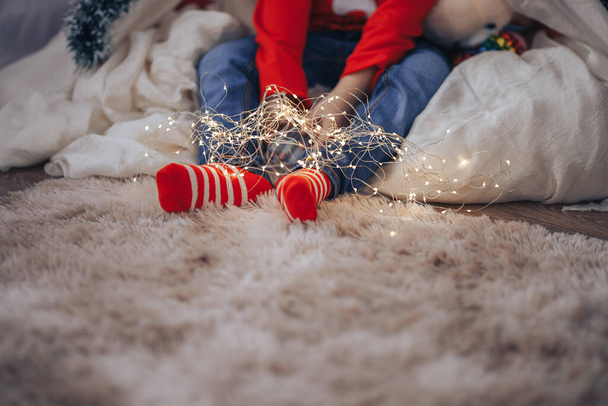 Criança em calças azuis e meias vermelhas segurando uma grinalda em suas mãos enquanto sentado em um travesseiro macio, Feliz Natal e Feliz Ano Novo
 - Foto, Imagem