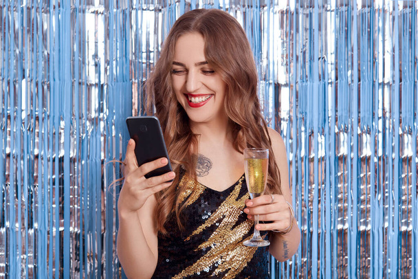 Happy smiling girl using smart phone and glass of wine in hands, model wearing stylish elegant black dress, looking at device's screen and laughing happily with glitter tinsel on background. - Foto, Bild