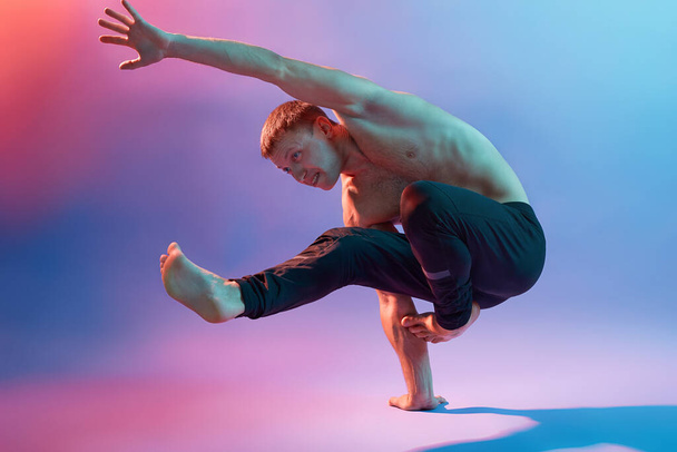 Acrobat showing his achievements, standing on one hand, demonstrating his power, gymnast training against neon background, guy wearing black pants. - Photo, Image