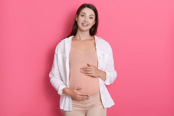 Young beautiful pregnant woman standing isolated over pink background and looking at camera with charming smile. keeping both hands on belly, has dark hair. - Foto, afbeelding