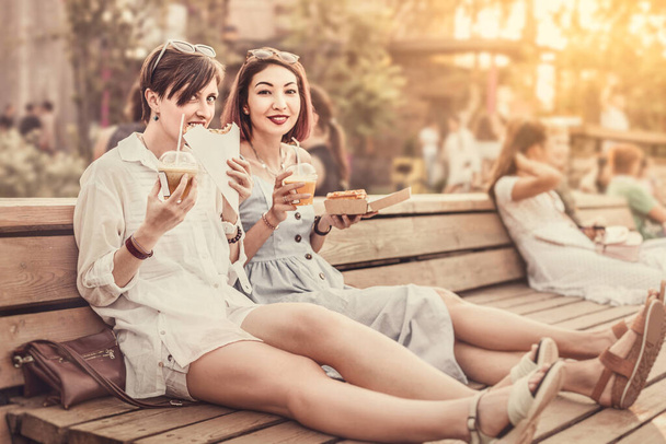 Kaksi onnellista naista ystäviä rentoutua puistossa ja syödä katuruokaa ottaa pois ja juoda virvoitusjuomia. Rento seurustelu ja kaupunkien pikaruokaa loma - Valokuva, kuva