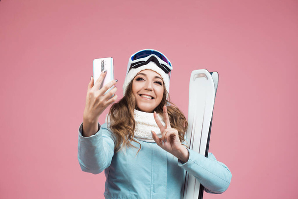 fun skier takes a selfie on her smartphone. A young woman with ski equipment in a jacket and hat, - Foto, Imagem