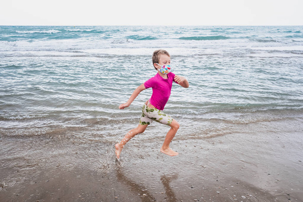 Chlapec běží podél břehu pláže v masce obličeje během nové normality. - Fotografie, Obrázek