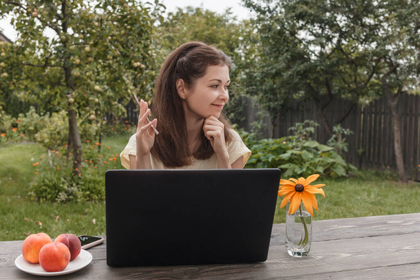 Fiatal nő egy könnyű pólóban távolról dolgozik egy laptop ül egy fa asztalnál a háttér zöld kert és nézi az oldalán - Fotó, kép