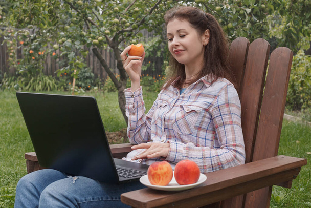 Una joven con una camisa trabaja remotamente en un portátil sentado en una silla en el jardín, comiendo un melocotón y pensando en algo - Foto, imagen
