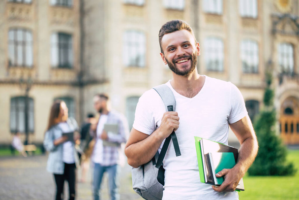 Young handsome bearded student guy in casual clothes with backpack and books in hands is walking and posing on a university  buildings background - Photo, Image