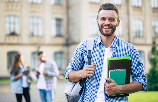 Young handsome bearded student guy in casual clothes with backpack and books in hands is walking and posing on a university  buildings background - Fotoğraf, Görsel