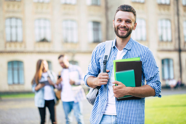 Young handsome bearded student guy in casual clothes with backpack and books in hands is walking and posing on a university  buildings background - Foto, imagen