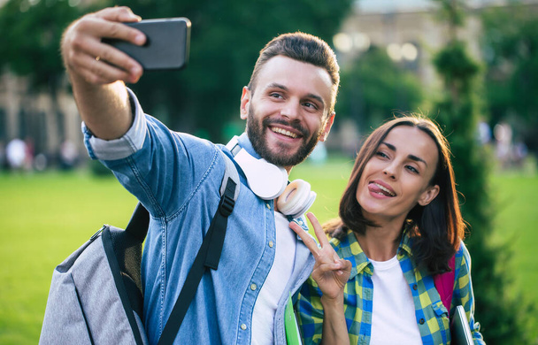 Beautiful young happy couple in casual clothes with backpacks are making selfie photo on smartphone - Φωτογραφία, εικόνα