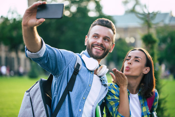 Beautiful young happy couple in casual clothes with backpacks are making selfie photo on smartphone - 写真・画像