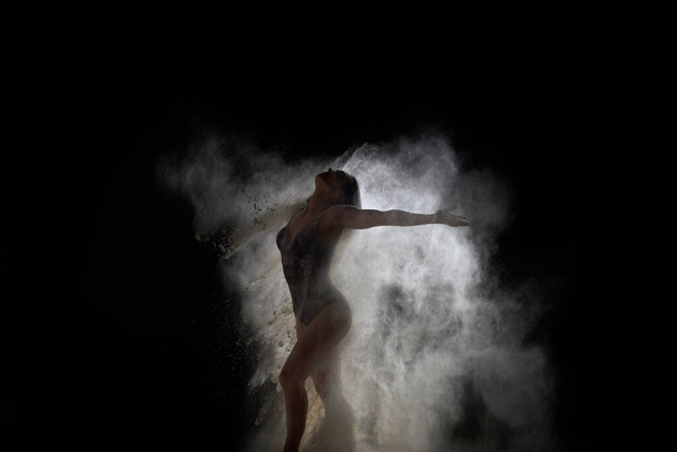 красива молода біла біла жінка в чорному костюмі зі спортивною фігурою танцює в білій хмарі борошна на чорному тлі, вибуху і вибуху в русі
 - Фото, зображення