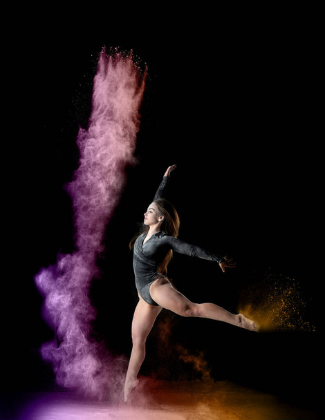 schöne junge kaukasische Frau im schwarzen Body mit einer Sportfigur, die in einer lila-gelben Mehlwolke auf schwarzem Hintergrund tanzt, Explosion und Ausdruck in Bewegung - Foto, Bild