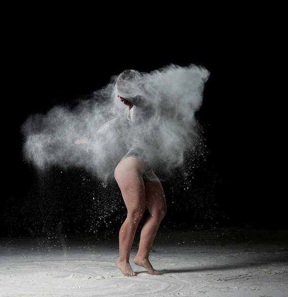 hermosa mujer caucásica joven en un body negro con una figura deportiva está bailando en una nube blanca de harina sobre un fondo negro - Foto, imagen